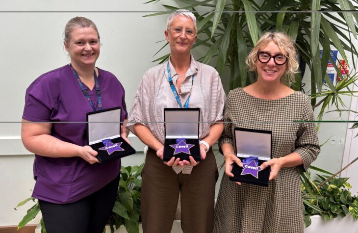 Three people stand in a line, smiling and holding small black boxes in front of them. Inside the boxes, there are purple star-shaped medals that read 'Cavell'.