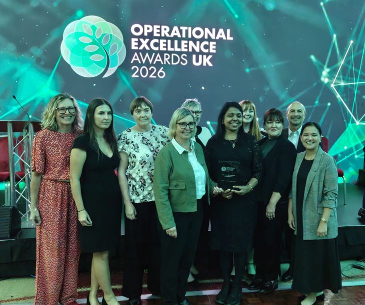 A large group of people stand in front of sign that reads 'Operational Excellence Awards UK 2026'. The smile as one person in the centre of the group holds an award. 