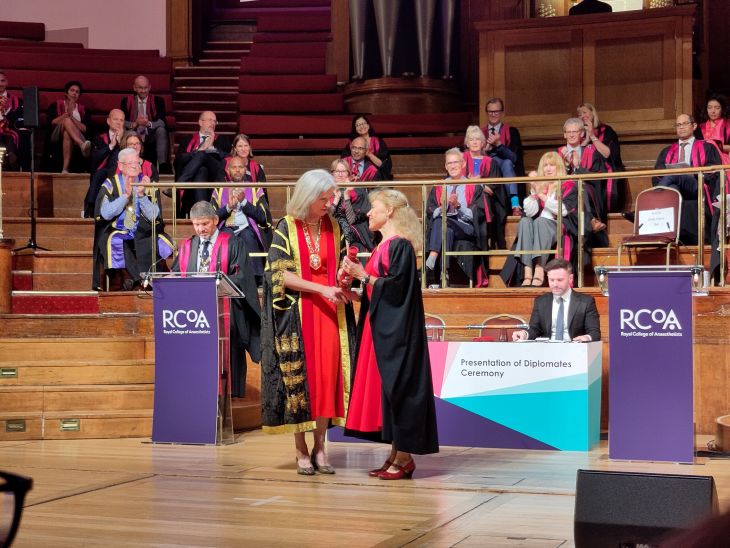 Two people in robes on stage. One is handing another a diploma, making an achievement.