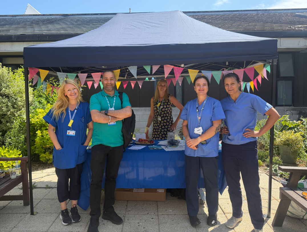 Group of people standing under a navy gazebo with colourful bunting. Three women and a man are wearing medical uniform and there is a woman in a patterned dress.
